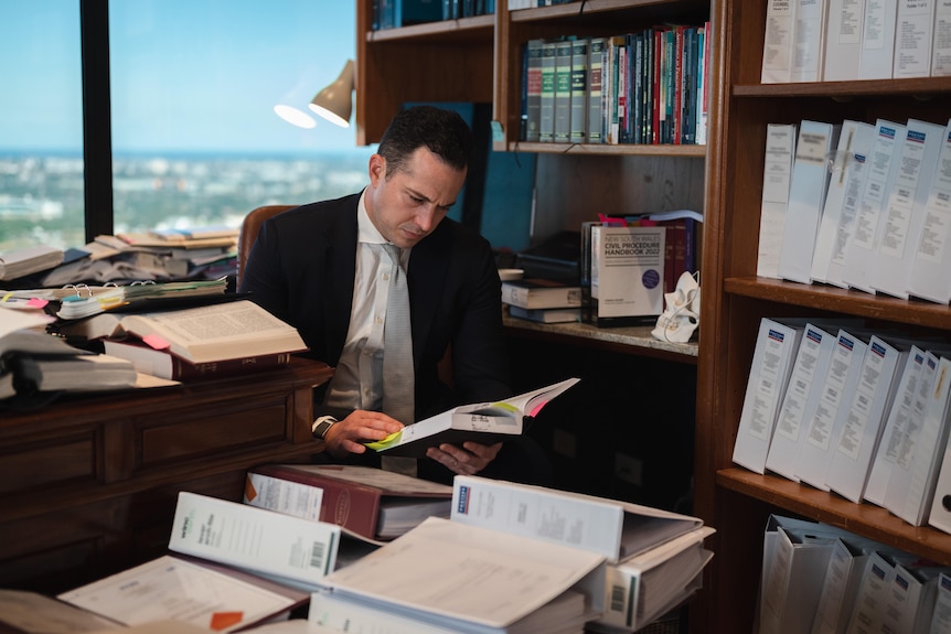 A man wearing a suit looks at a book, surrounded by folders in his office. There is a view of Sydney in the background.