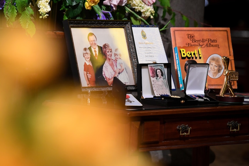Photographs of Bert Newton and a gold Logie on display with floral arrangements in the background.