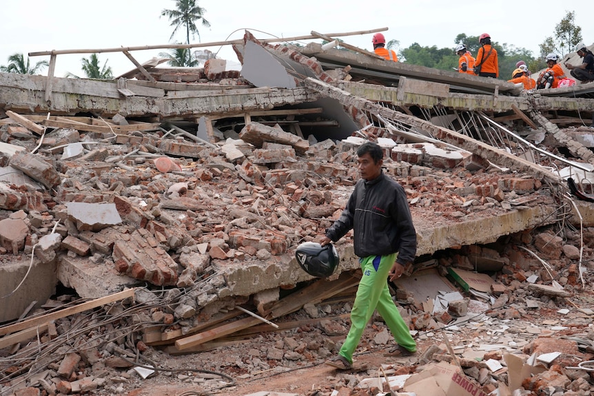 a man walks in sandals past a completely collapsed building. rescue workers can be seen in the background