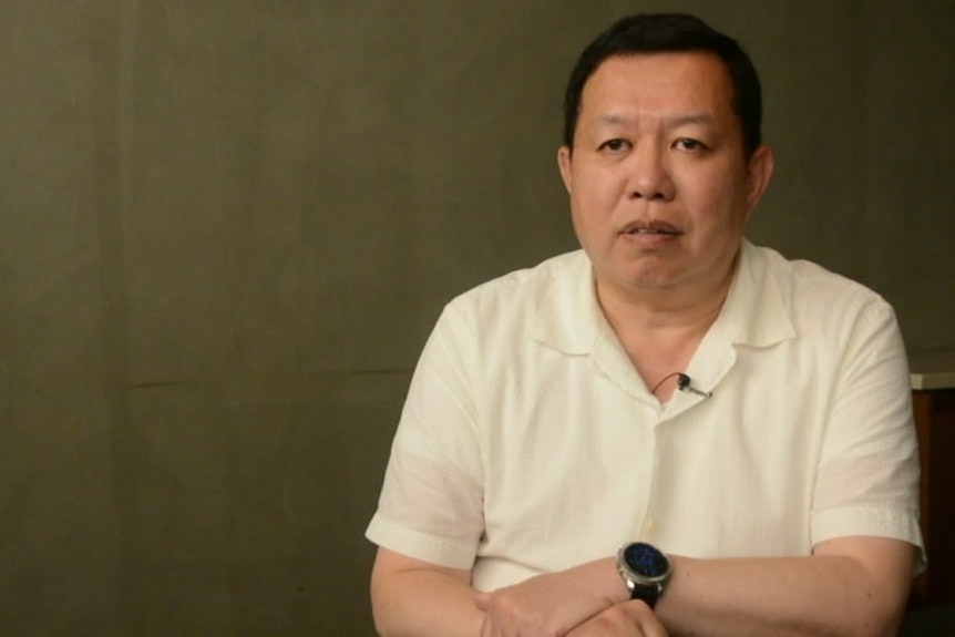 A South Korean man sits with his arms crossed at the wrist wearing a watch and a cream collared shirt.