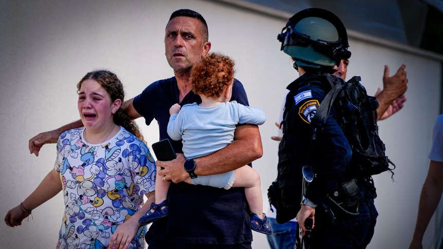 A man holding a baby and a crying girl walk past a police officer in a helmet 