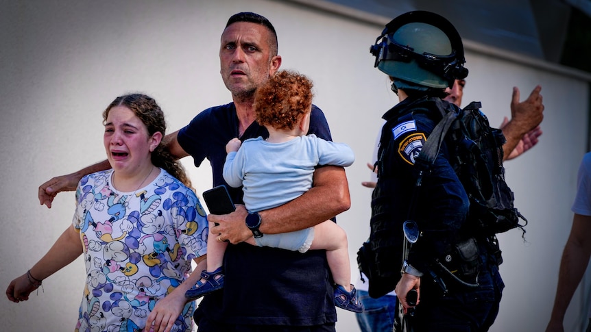 A man holding a baby and a crying girl walk past a police officer in a helmet 