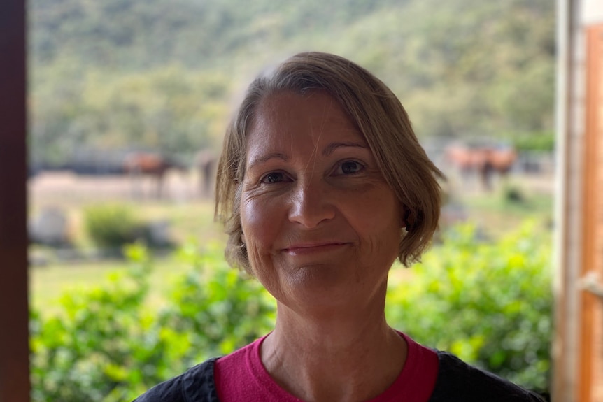 A fair-haired woman in a blue and pink top standing in front of a window or doorway to a garden-like backdrop