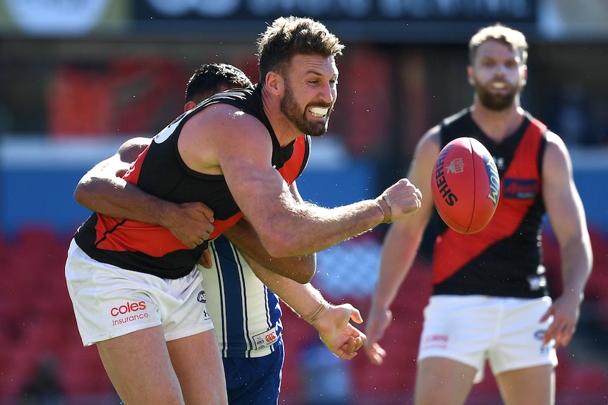 An Essendon AFL player gets a handball away while being tackled by a North Melbourne opponent.