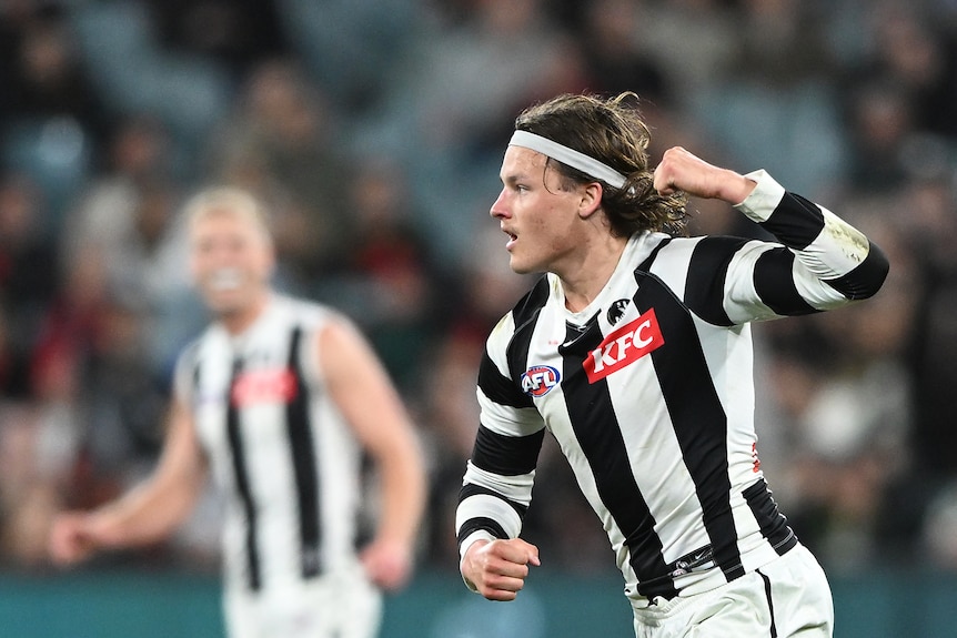 A male AFL player raises his fist in celebration, wearing a black and white guernsey, as a teammate celebrates behind him.