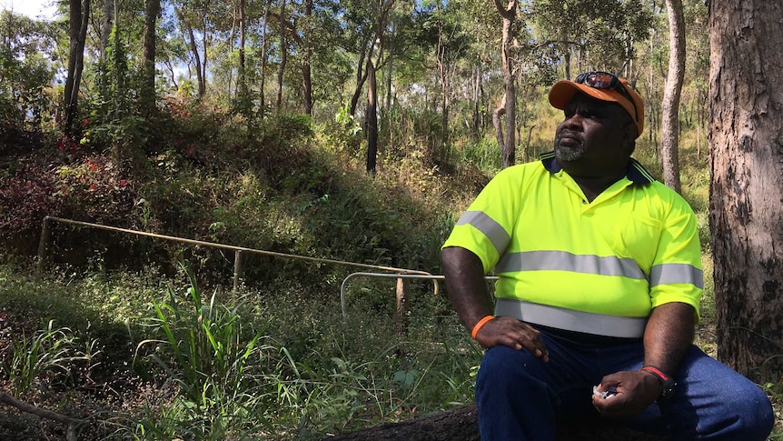 An Indigenous man sits on a large log looking at the bush around him in a site cleared by rangers