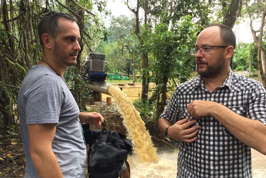 Marsic and Willacy talking with torrent of muddy water pouring out of pipe in background.