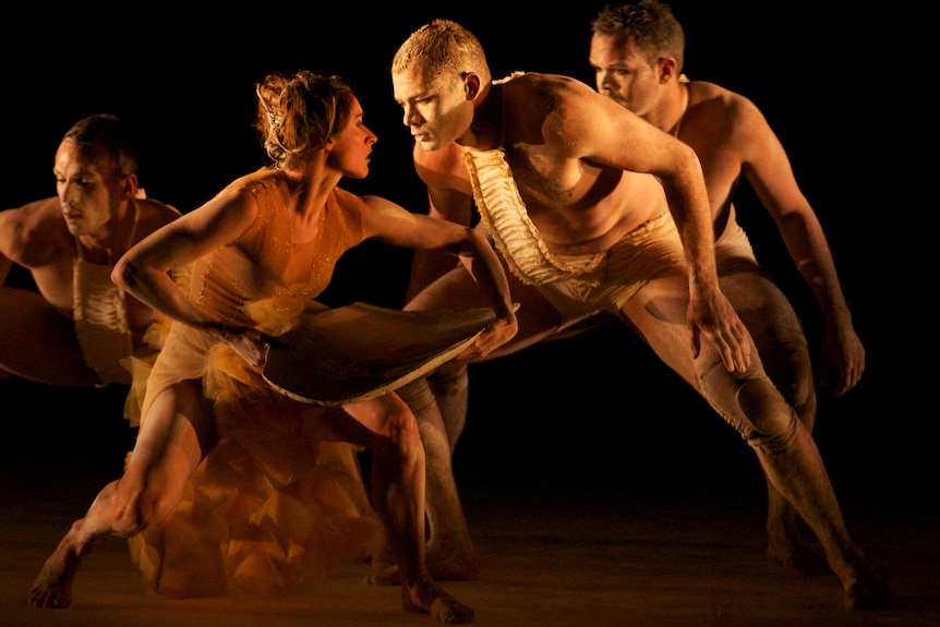 A painted up female and male dancer stare intensely at each other on stage near other others.