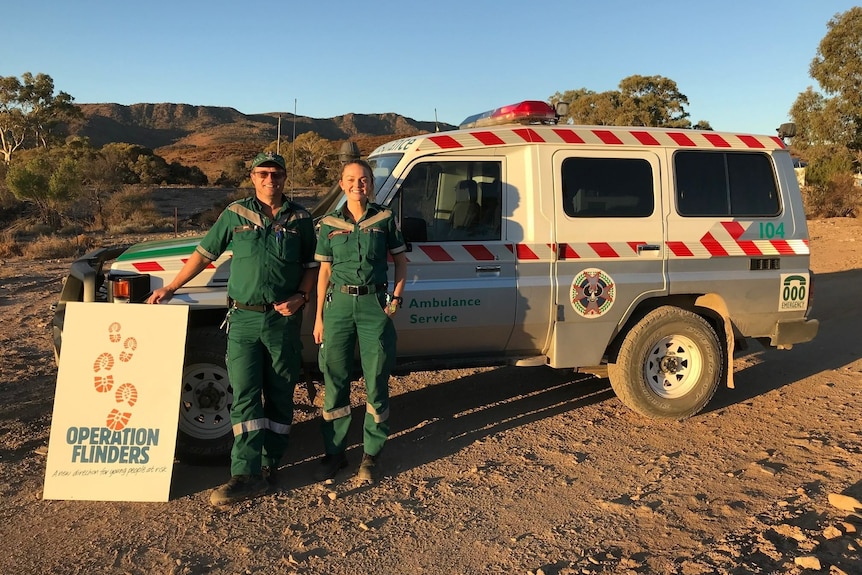 A man and woman wearing a green ambulance uniform stand in front of a four wheel drive vehicle. 