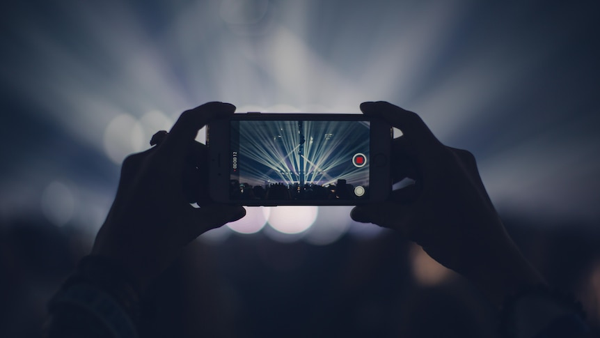 A crowd member holds their smartphone with two hands, recording. The stage is seen through the screen. 