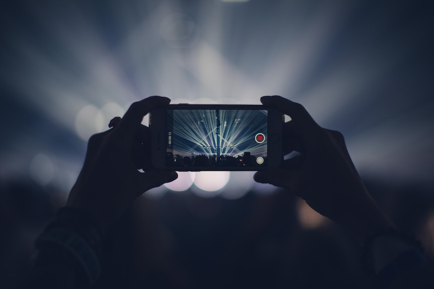 A crowd member holds their smartphone with two hands, recording. The stage is seen through the screen. 