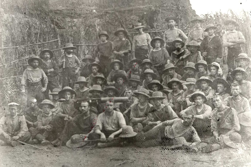 A few dozen naval men stand in front of a thatched building, what appears to be a machine gun sits among them.