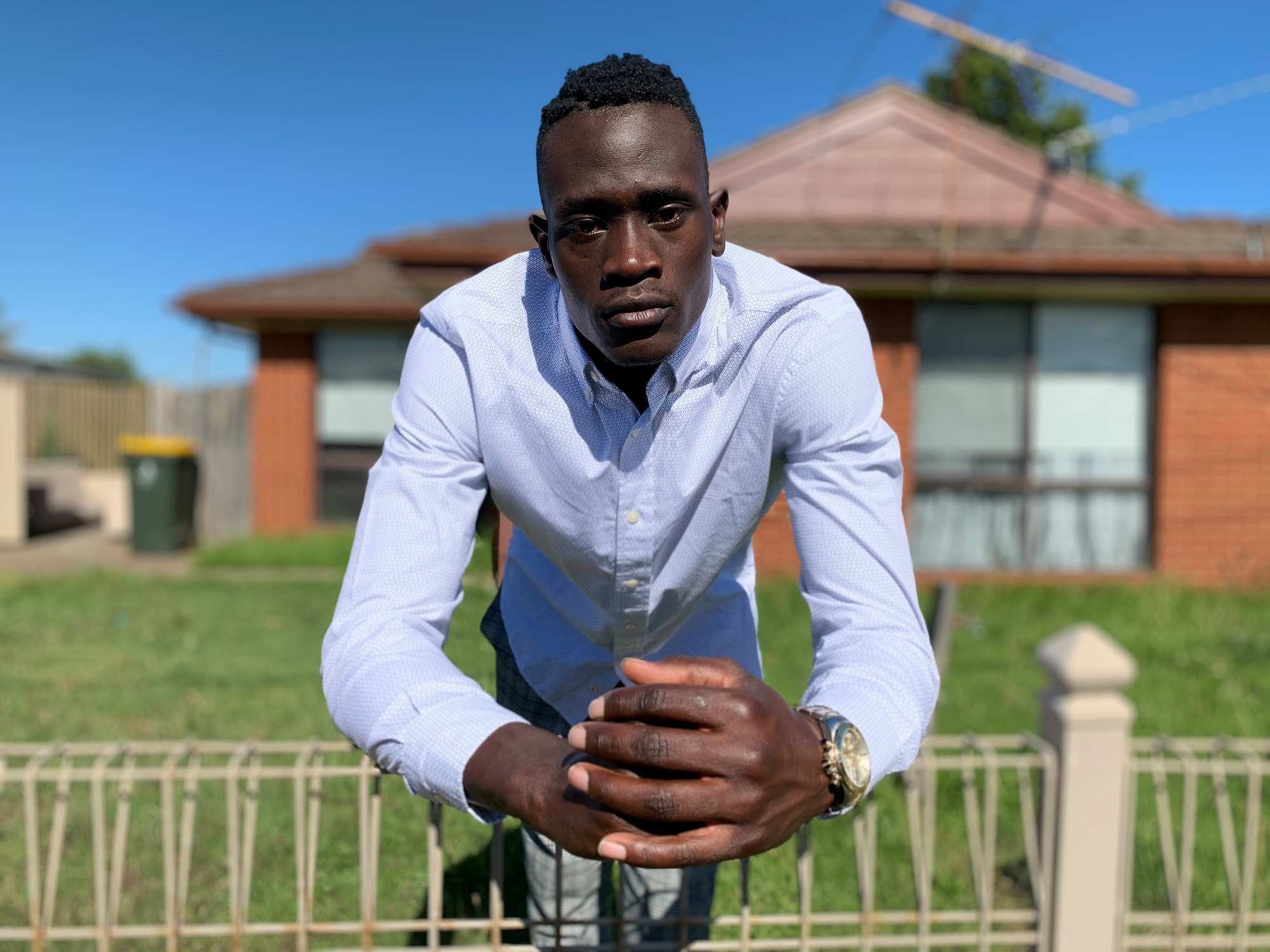 A young man leans against a fence with his arms looking into the camera.
