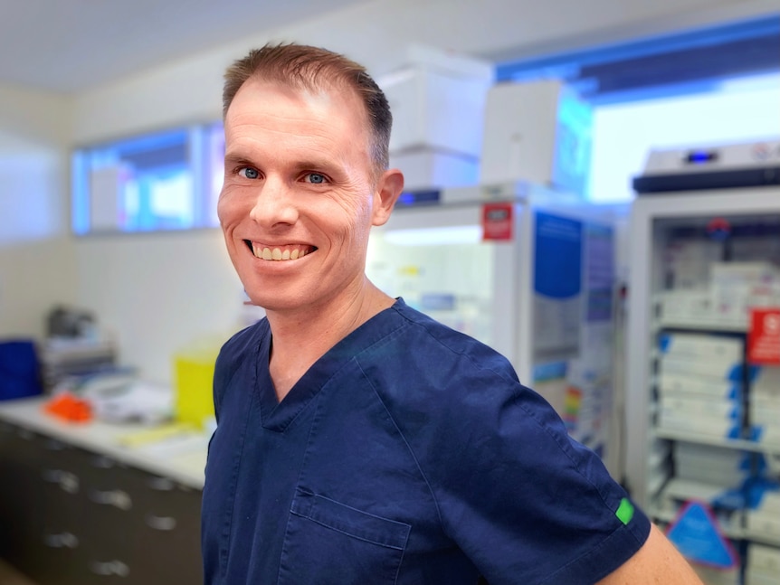 A man in blue scrubs smiles at the camera in front of medical fridges.