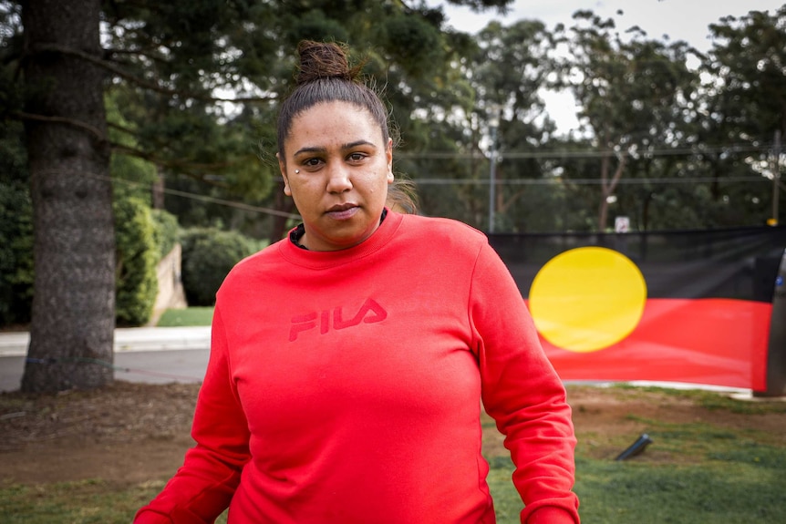 An Indigenous woman in a bun and red jumper, an Aboriginal flag behind her