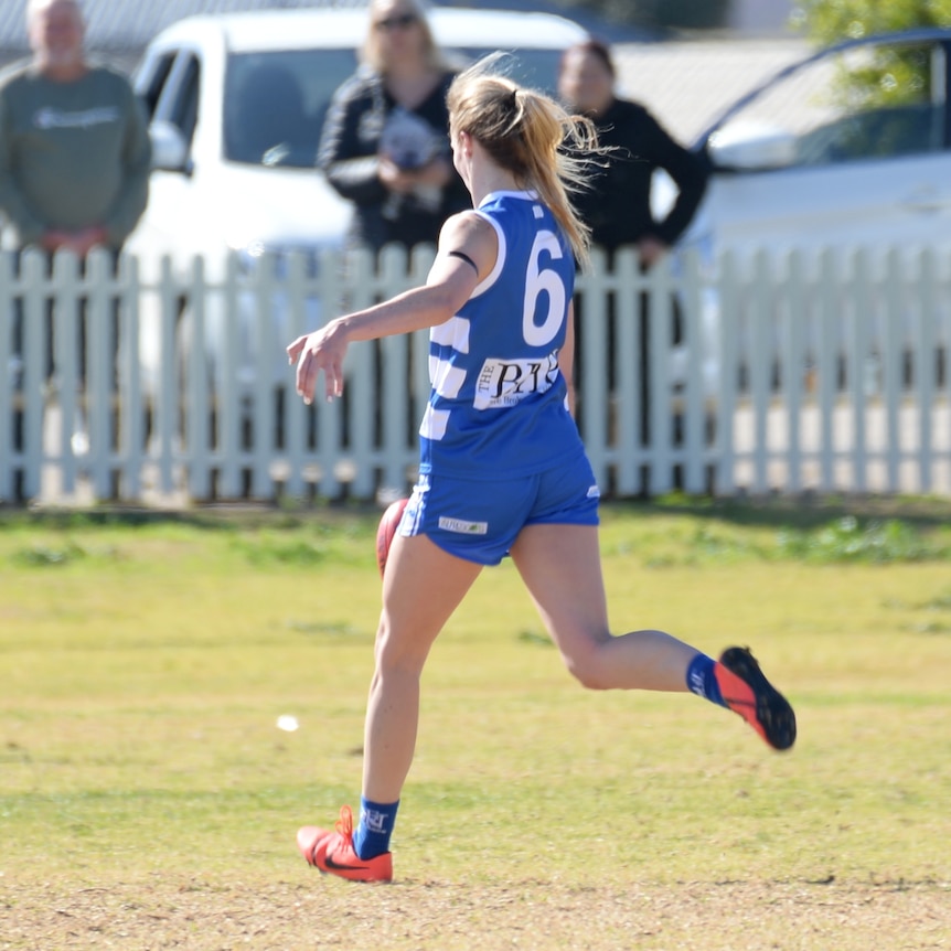 A blonde haired woman wearing a blue AFL uniform kicks a red football at a match in Broken Hill. 
