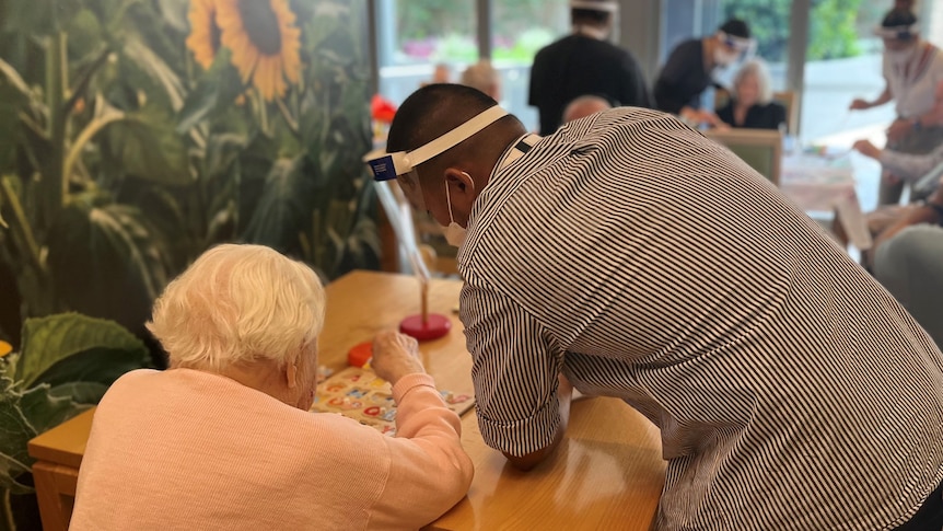 An elderly woman seen from behind sits at a table with a puzzle, with a male nurses leaning on the table.