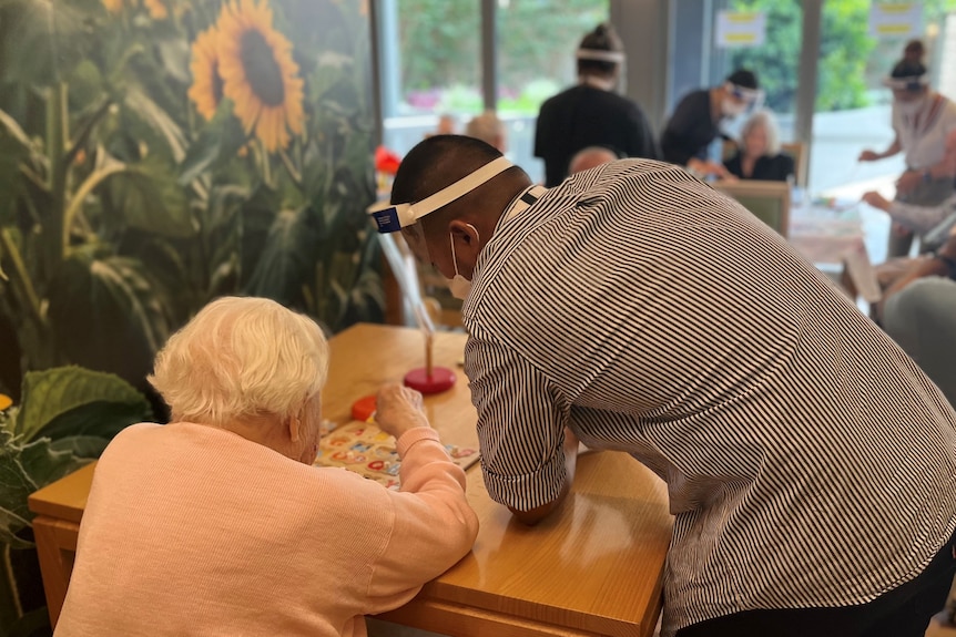 An elderly woman seen from behind sits at a table with a puzzle, with a male nurses leaning on the table.