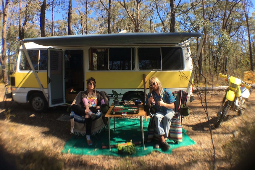 A young woman and man sit in front of a yellow and white minibus, with an awning. Tall gum trees are in the background.