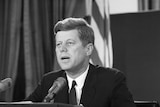Black and white photograph of John F Kennedy behind a large desk in the Oval Office addressing several journalists and cameras
