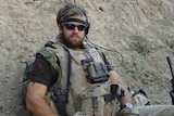 A man wearing a camouflage vest leans against a dirt ditch. 