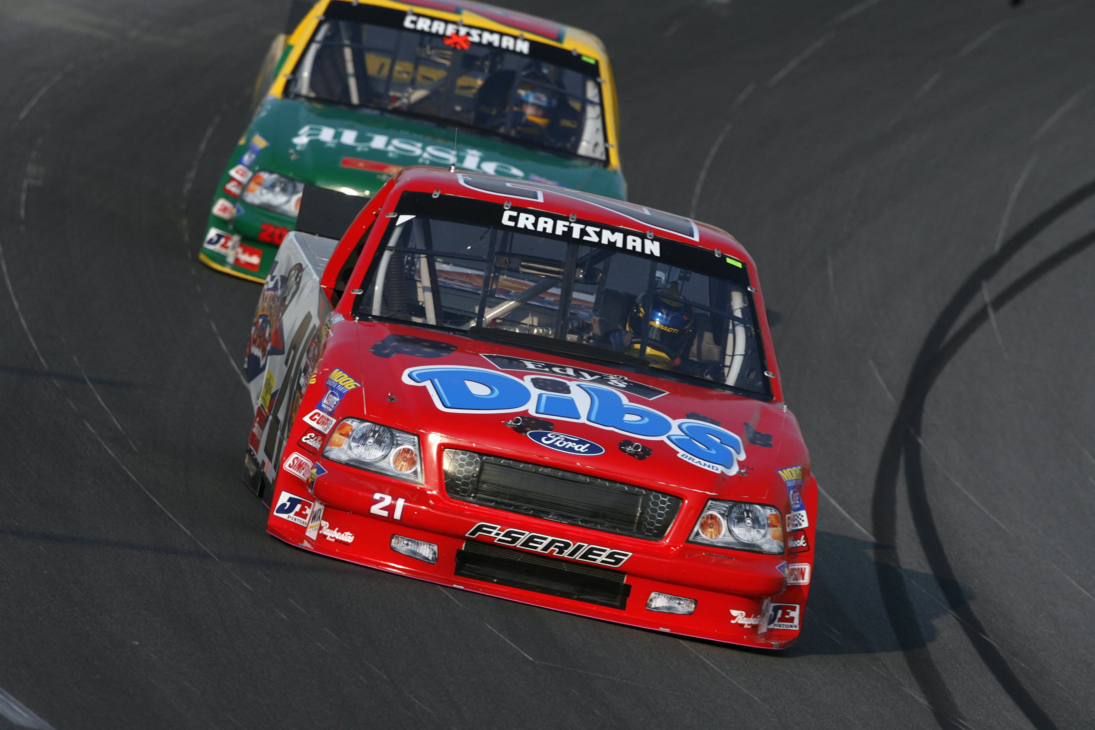 two NASCAR cars driving on a NASCAR track, in red and one in yellow and green