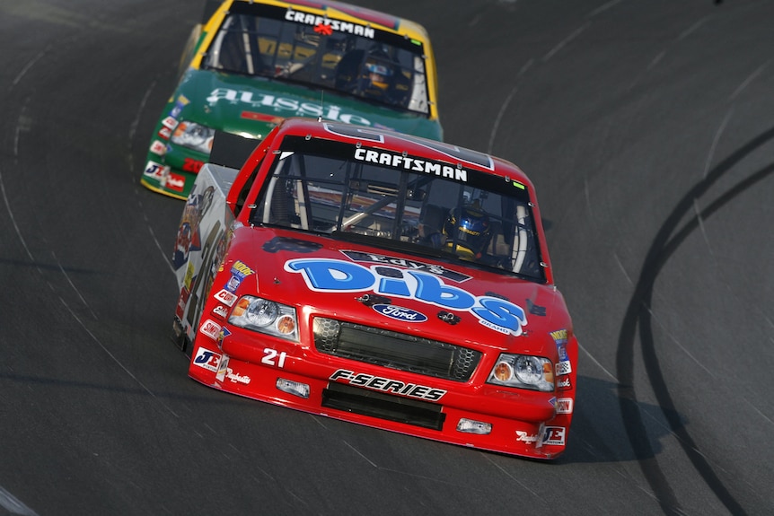 two NASCAR cars drive around a NASCAR track, on in red and one in yellow and green