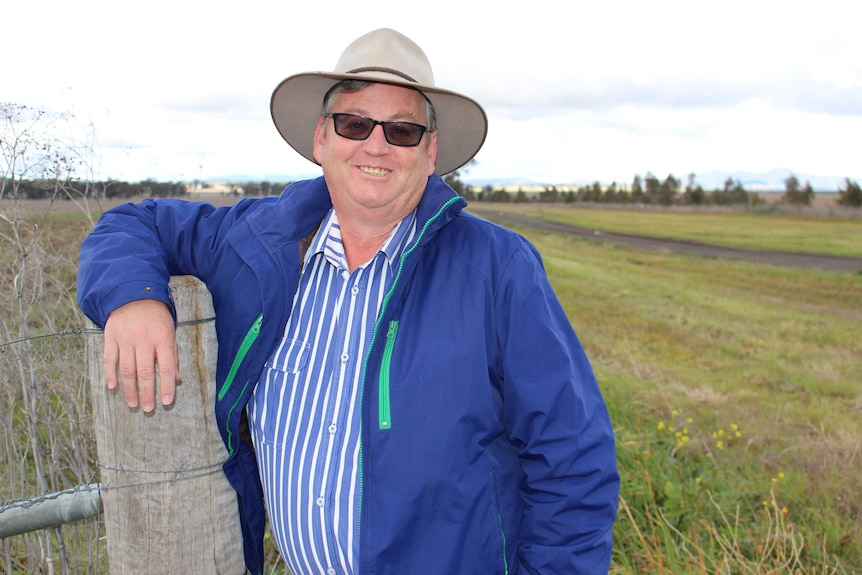 A man stands smiling in a paddock, resting his arm over a fence strainer post