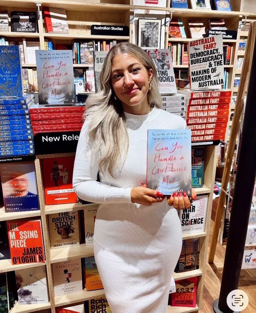 Bek wears a white figure-hugging dress blonde hair, smiles, holds a book, book shelves behind her.