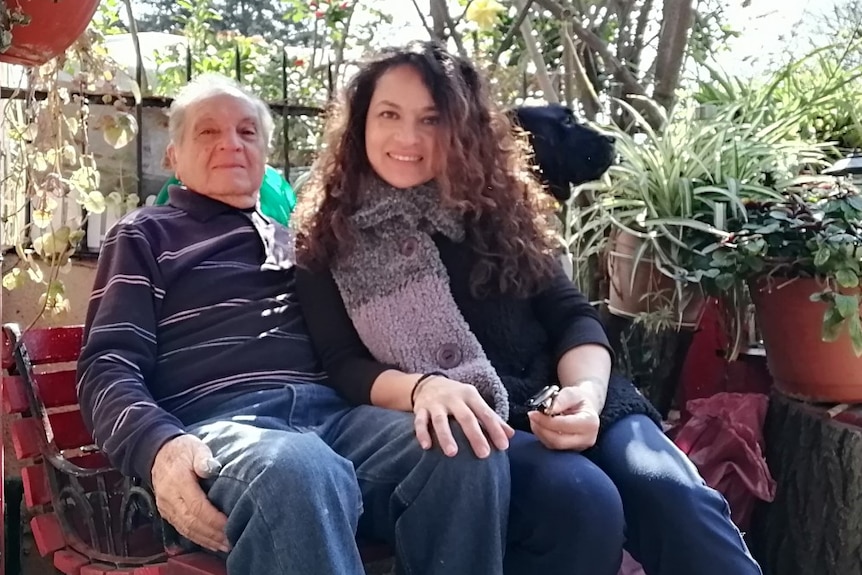 An elderly man and his daughter sit on a bench in a garden.