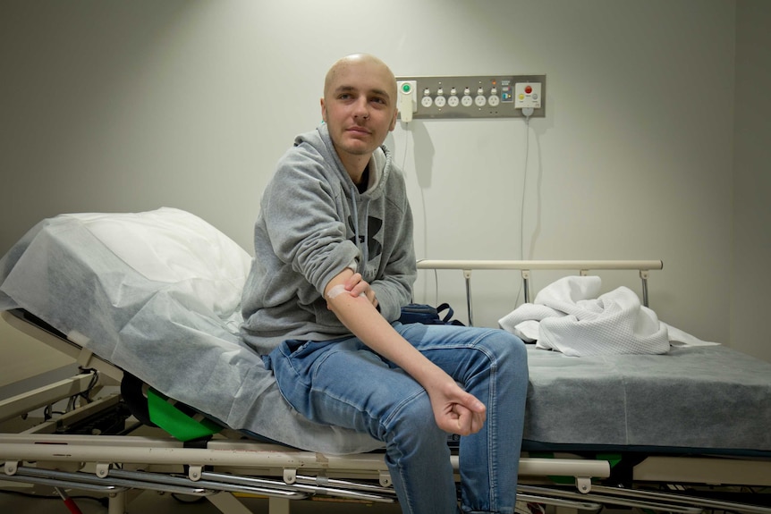 Josh Dickinson sits on a hospital bed holding his arm where he's just received an intravenous injection.