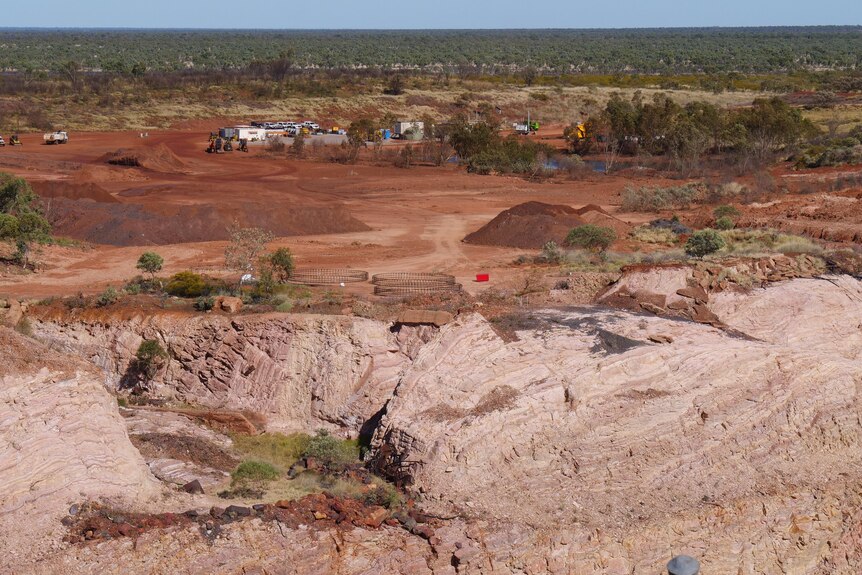 A view looking down at an open cut mine in red earth, with trucks, graders and sheds in the background.