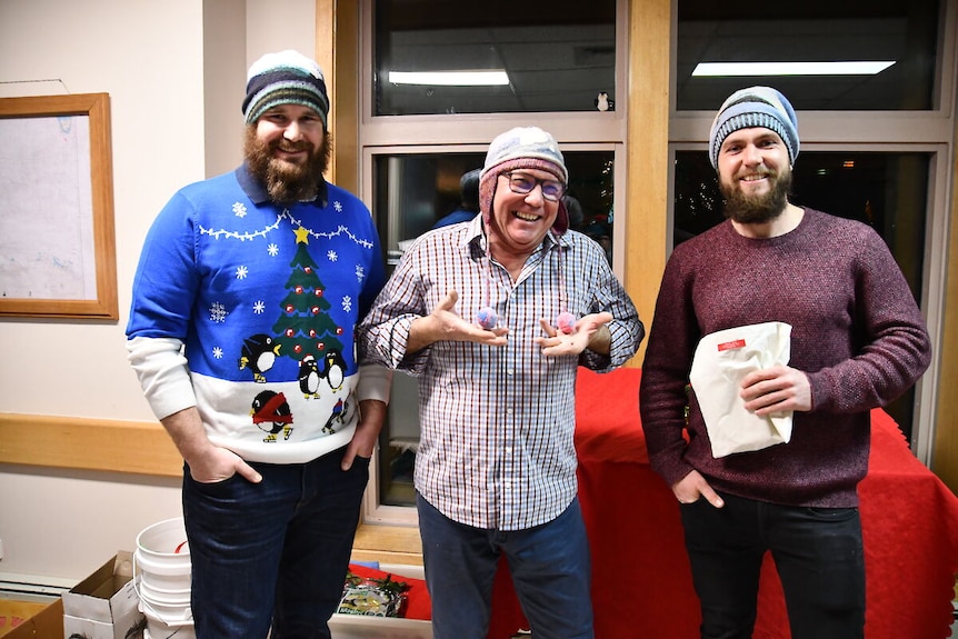 Three men stand inside smiling, wearing patterned beanies and long-sleeve tops.
