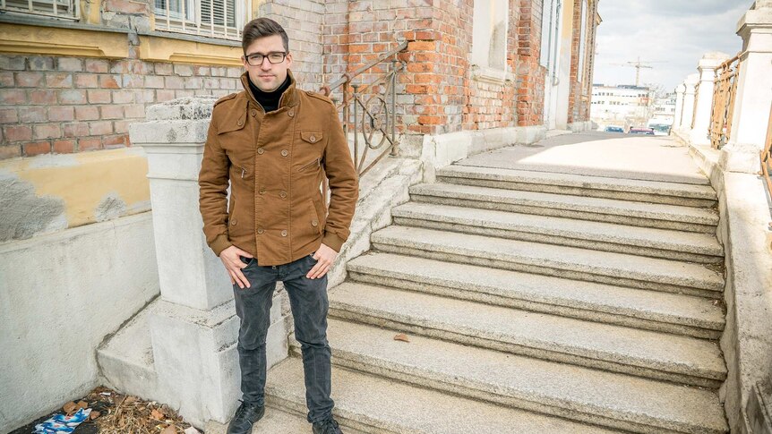 A young man wearing glasses and a heavy coat stands in front of a building.