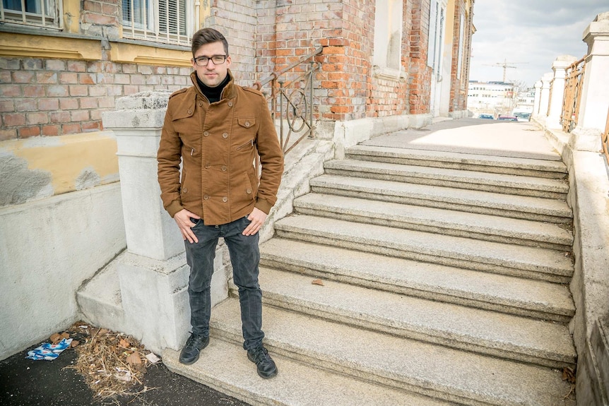 A young man wearing glasses and a heavy coat stands in front of a building.