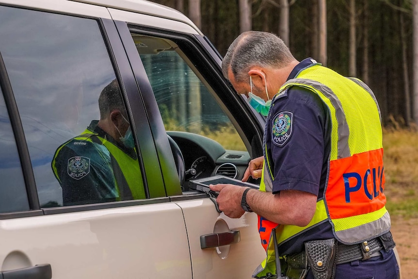 A man in police uniform, a high vis vest and face mask leans down to an open window of a driver's seat, a tablet in hand.