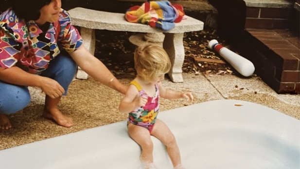 a little girl with blonde hair and a multi coloured suit is being held up by the pool by an older woman with dark curly hair