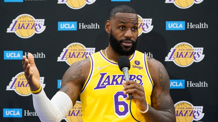 LeBron James, in his Los Angeles Lakers jersey, gestures while holding a microphone and sitting at a podium at NBA media day.