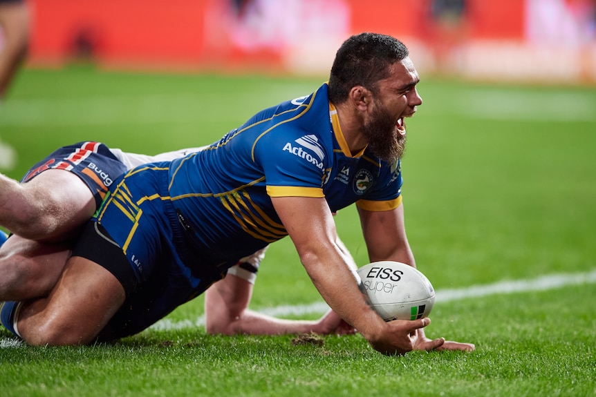 Isaiah Papali'i lies on the floor lifting his torso off the ground with his mouth wide open in a grin while holding a rugby ball