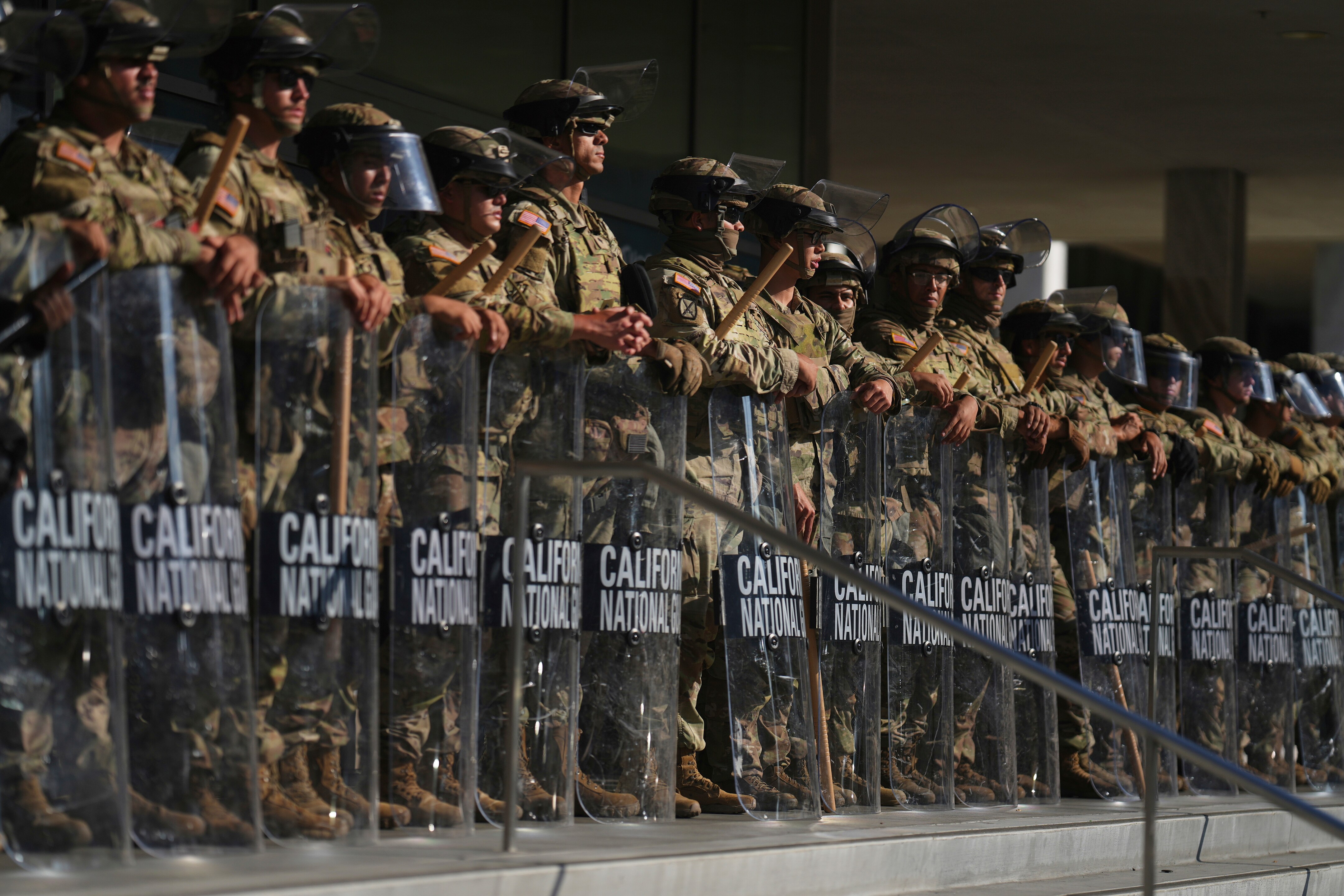 A line of soldiers in camouflage gear holding riot shields on the steps of a federal building.