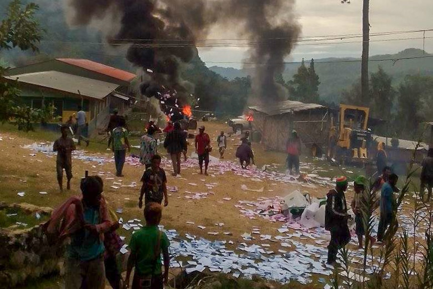A small PNG village, with several buildings on fire and pink and blue papers strewn across the ground 