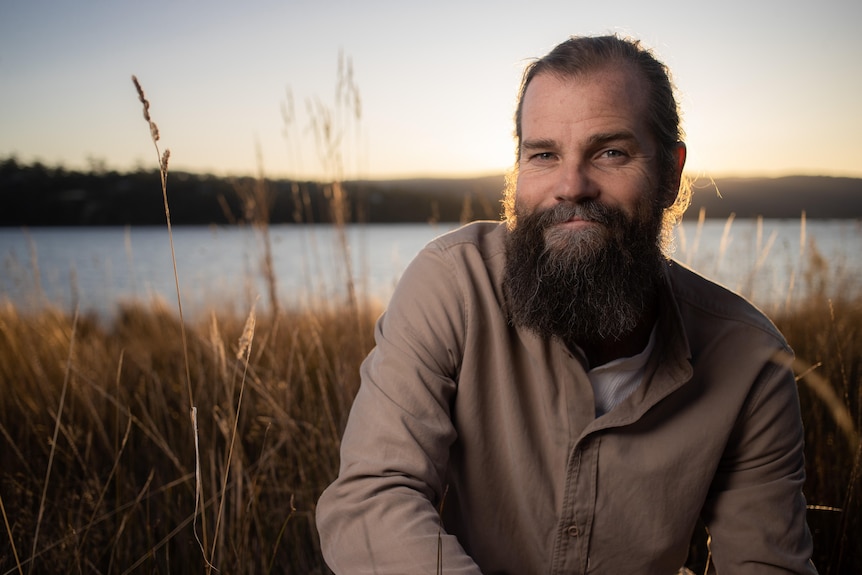 A bearded man smiles slightly in front of lake and reeds of grass at sunset