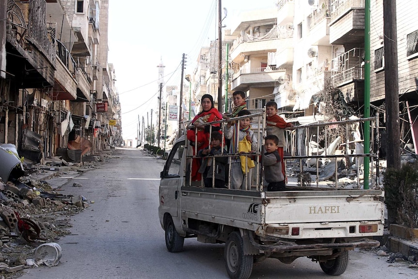 A family are crowded onto the back of a truck in the middle of street blown apart by bombing.