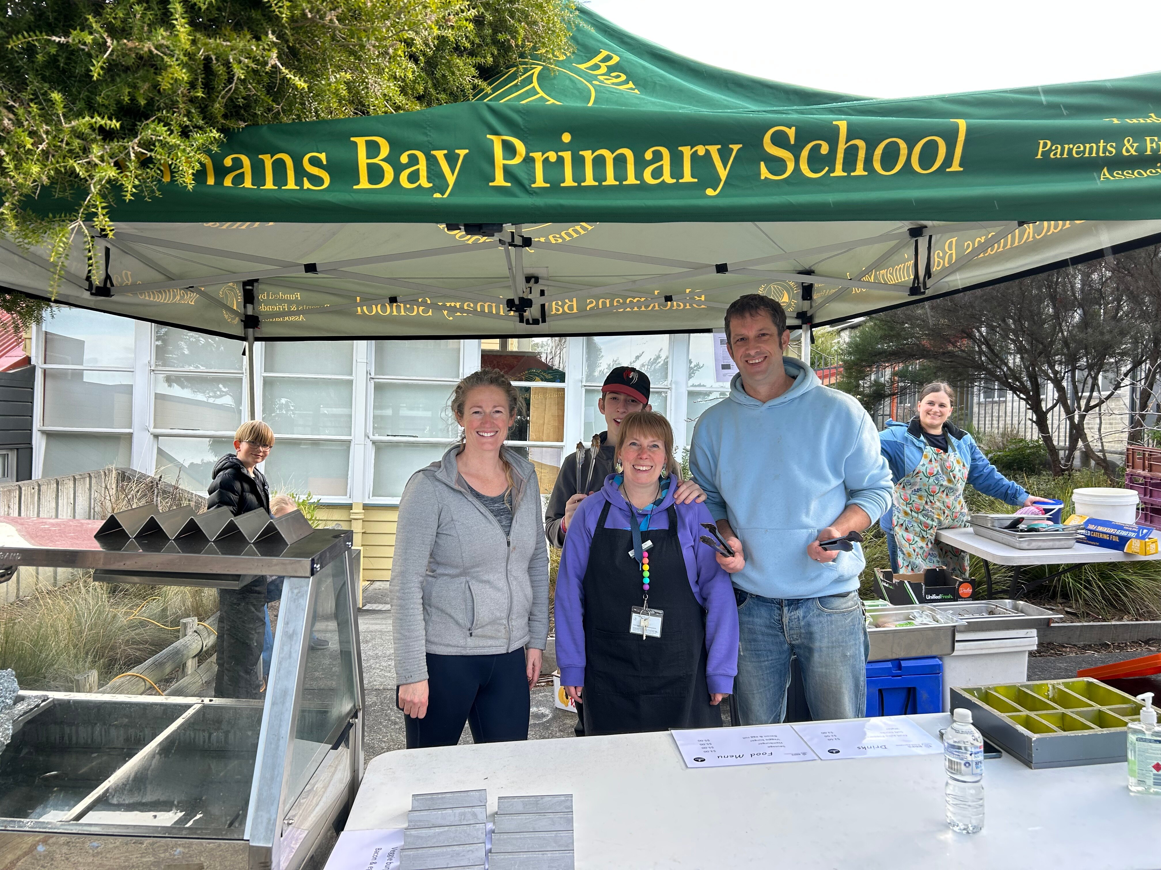 A family stand in front of a barbecue, underneath a school marquee.