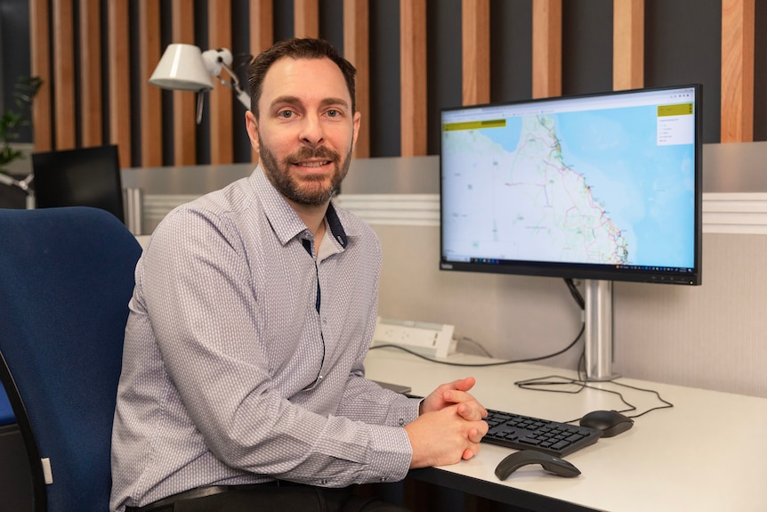A slightly smiling man in business clothes in front of a computer with a map of Queensland, has neat black beard and moustache.