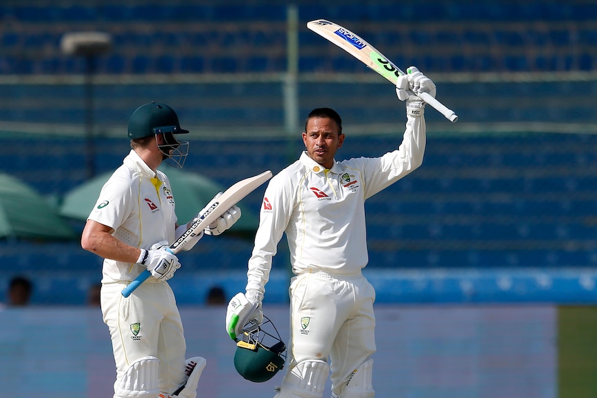 Usman Khawaja removes his helmet and holds his bat up while standing next to Steve Smith