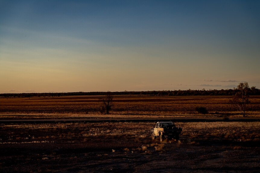 A white ute is facing a vast expanse of dry, red property, basked in warm evening light as the sun sets.