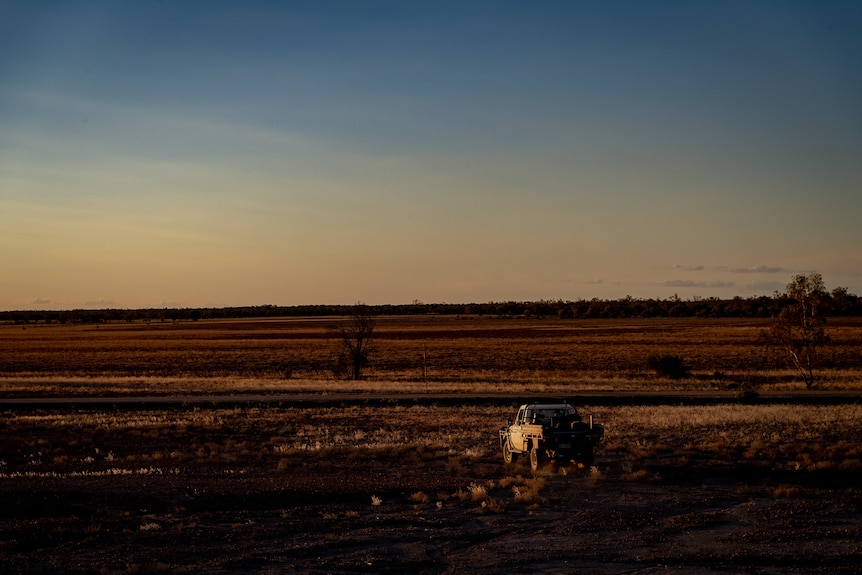 A white ute is facing a vast expanse of dry, red property, basked in warm evening light as the sun sets.