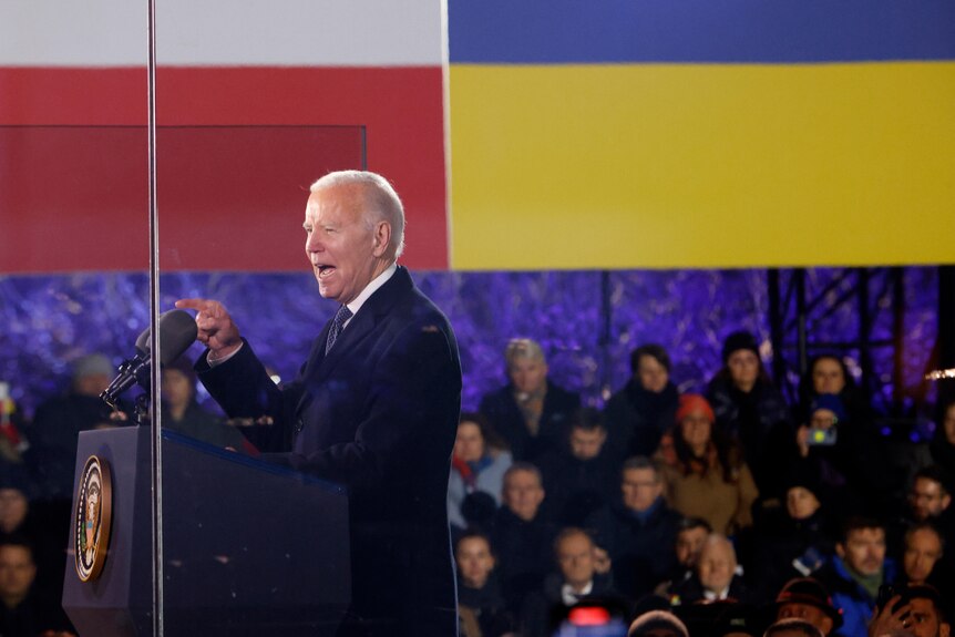 An elderly man in a dark suit points as he speaks from behind a lecturn, with Ukrainian and Polish flags hung behind.