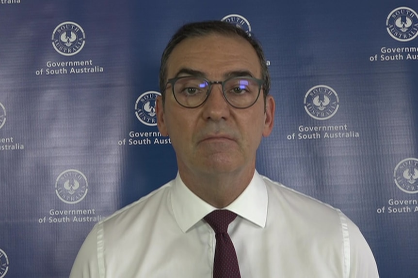 A man wearing black glasses, a white shirt and red tie stands in front of a blue background